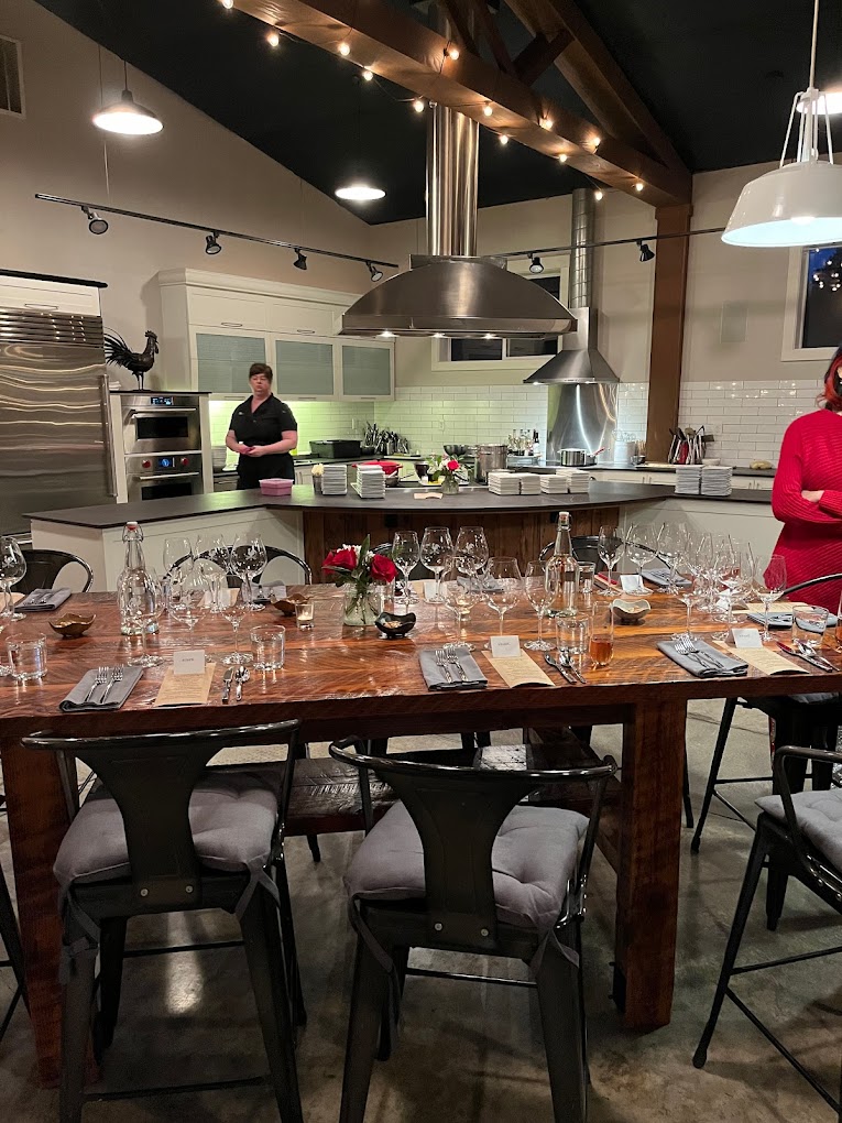 The interior of The Kitchen At Middleground Farms. There's a kitchen and a large wood table set up with wine glasses, cutlery, and modern metal chairs.