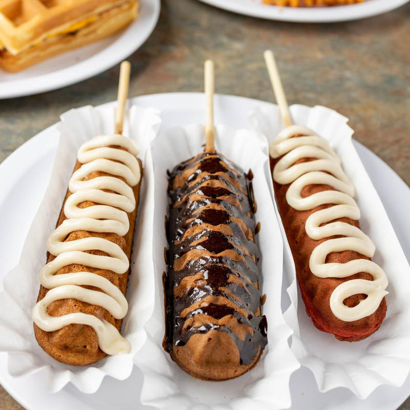 Three Lolly Waffles lined up. It's a waffle in the shape of a corndog on a stick, drizzled in sweet sauce.