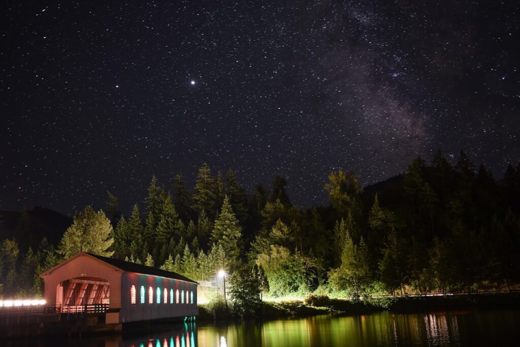 Stars hang in the night sky above the Lowell Covered Bridge, with trees in the background. Colorful lights come out from the bridge openings and light up the night.