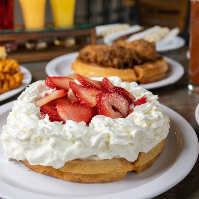 A waffle with whip cream and sliced strawberries on top. In the background there's a large waffle with fried chicken on top.