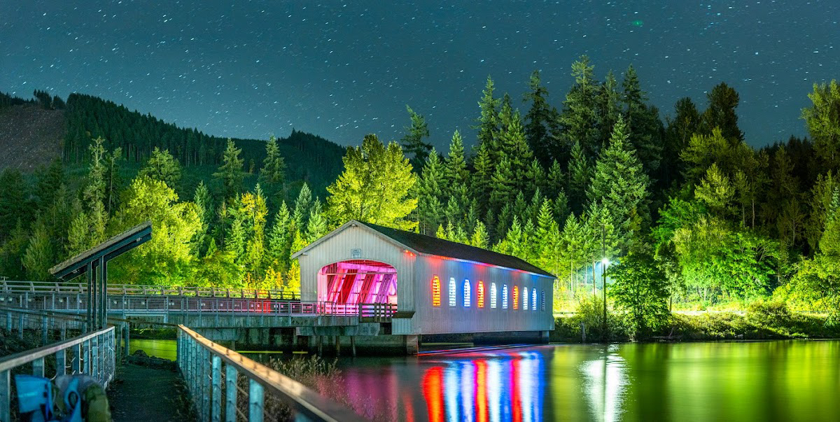 A nighttime shot of the Lowell Covered Bridge. The bridge is white and has many colorful lights coming out from inside it. Stars hang in the sky, nad green water sits below. Trees stand tall above the bridge in the background as a path leads to it in the foreground.