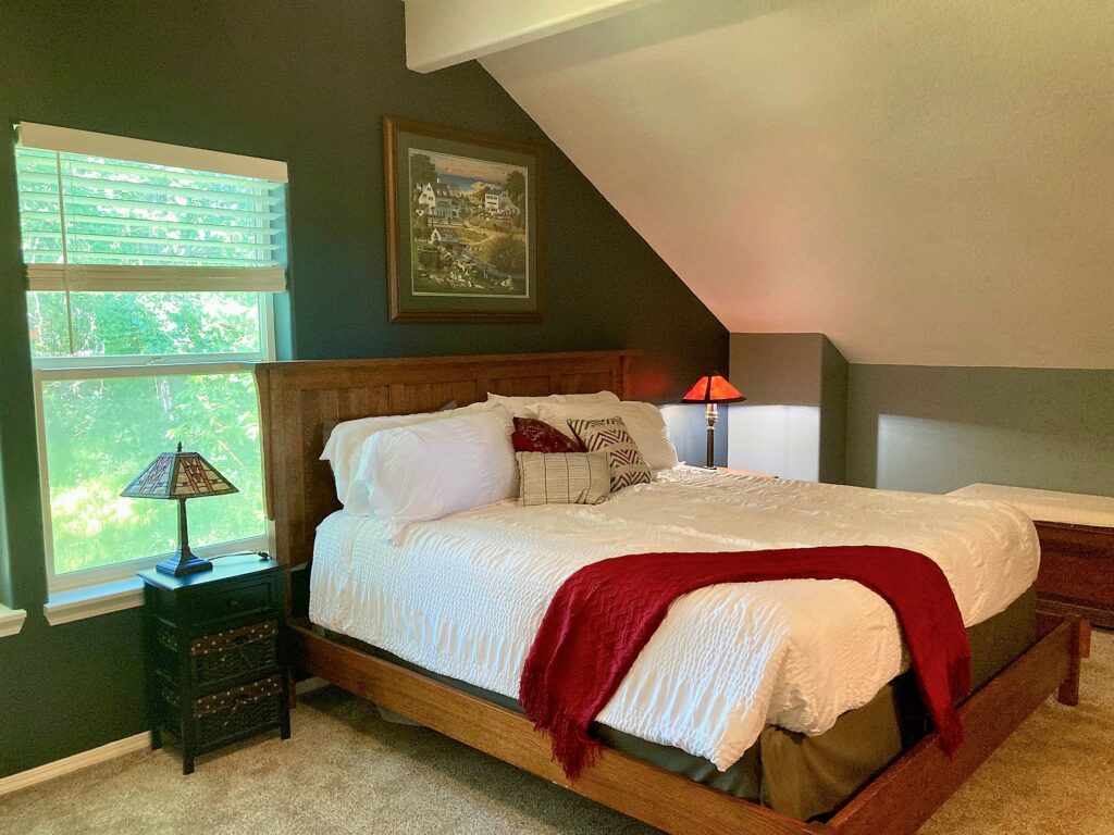 The King Room at the Owl's Nest Inn Bed And Breakfast. A large bed with white blankets and a red blanket draped over one corner. The room is dark green, and white and has a calming feel to it.