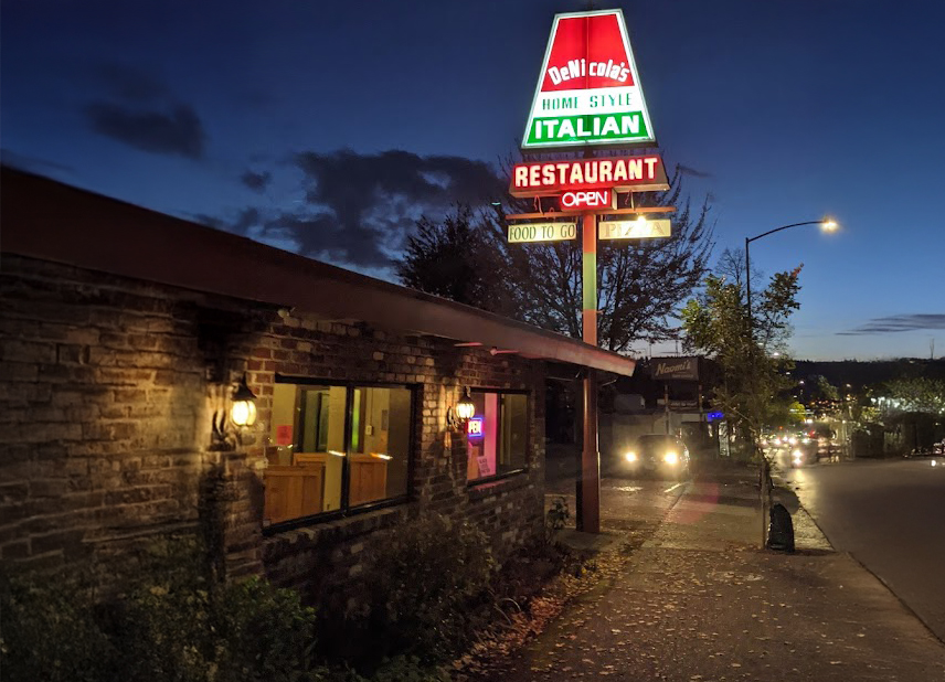 The outside of DeNicola's restaurant at night. A large red, white, and green sign lights up the area proclaiming: 'DeNicola's Home Style Italian Restaurant, Open, Food To Go, Pizza.'