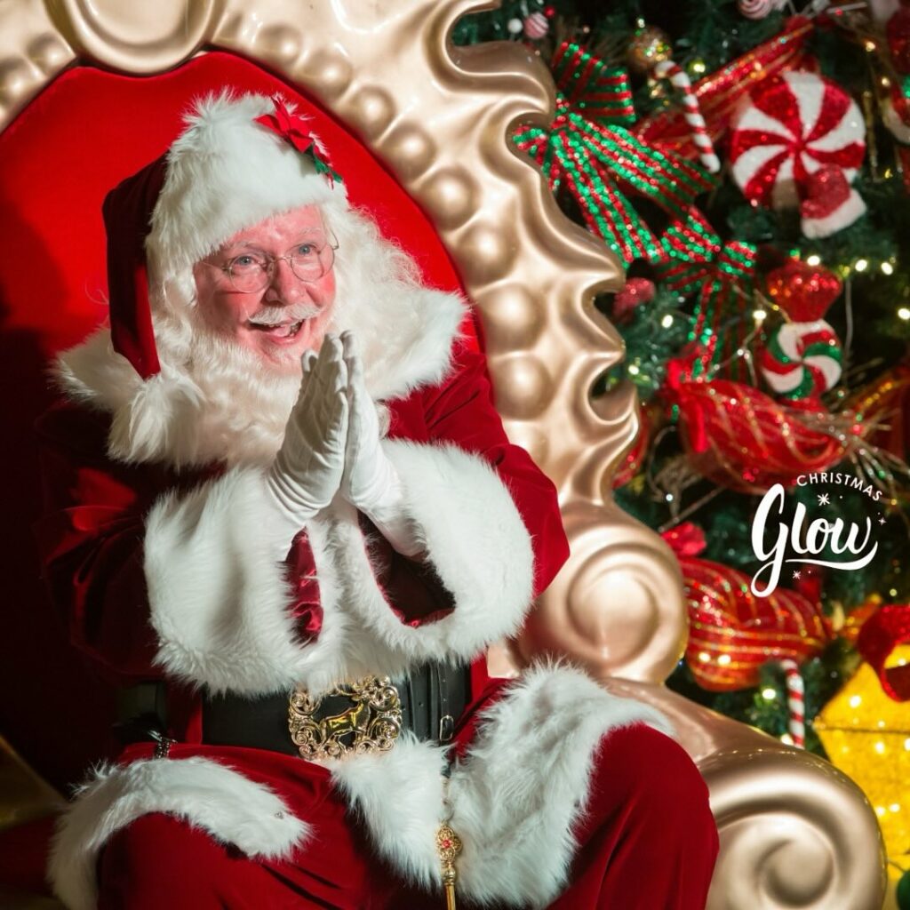 Santa Clause sits in a richly decorated red and bronze throne with his hands held together, looking excited to greet children.