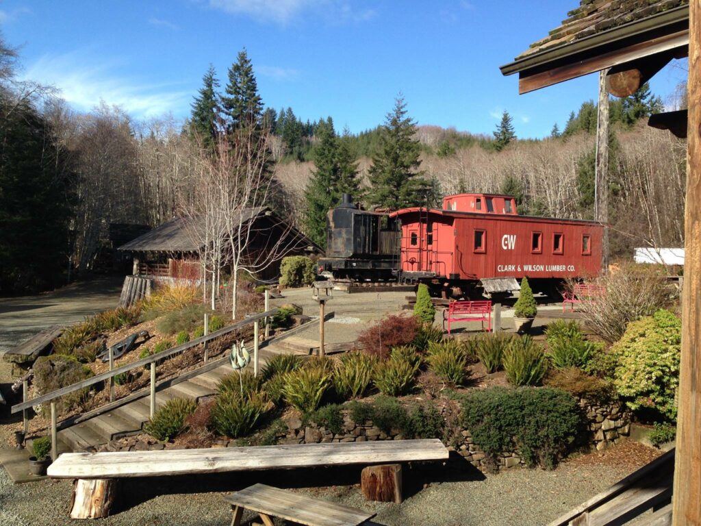 The Camp 18 museum on a sunny day. There's a red train car, and stairs leading up to several other things to see. The grounds have bushes and other landscaping, and a forest sits behind the museum.