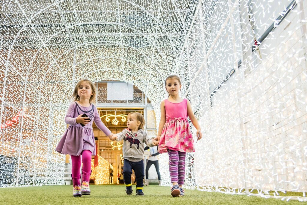 Three young children walk through a large white light tunnel created from Christmas lights.