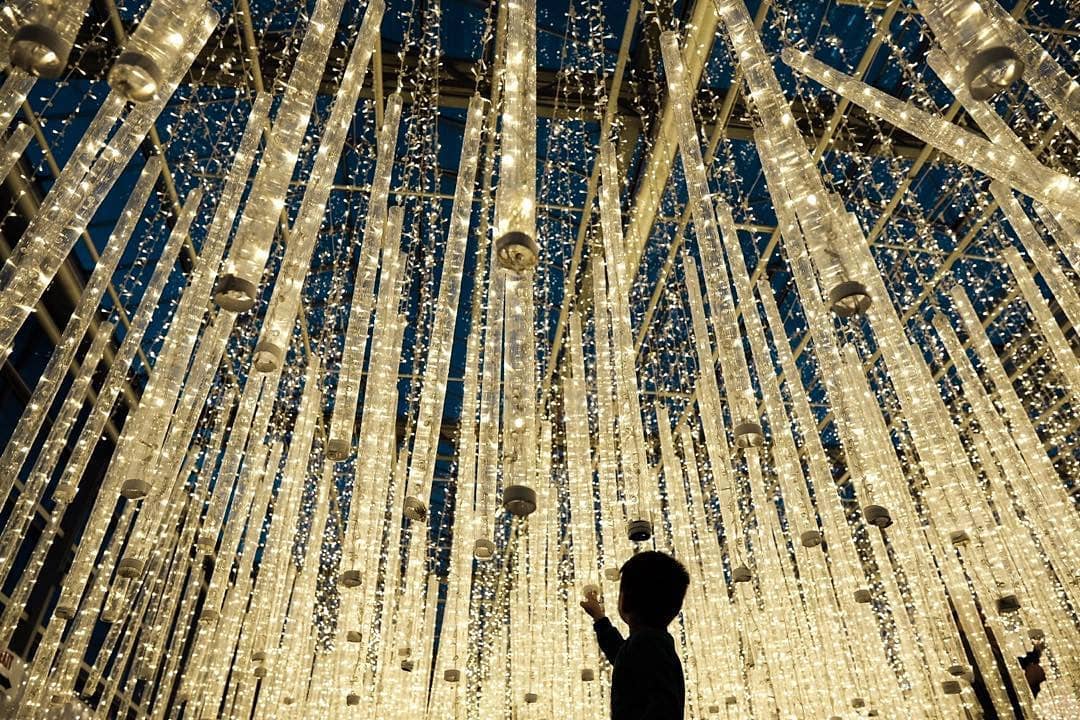 The silhouette of a small child stares up at dazzling white lights that hang down from the ceiling.