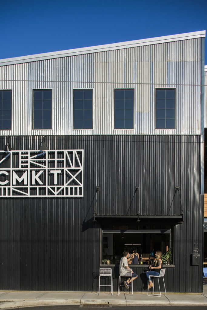 The outside of Mac Market. It's a black and tan metal warehouse. In this photo, people sit at a walk up window bar in the side of the building.