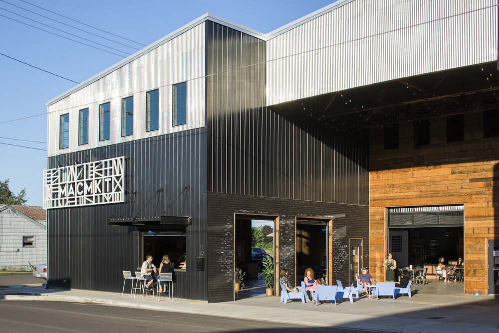 The outside of Mac Market. It's a black and tan metal warehouse. In this photo, people sit at a walk up window bar in the side of the building, and just around the corner there's a large outdoor seating space, which is partially covered. Metal and warm wood tones make it have such a friendly vibe.