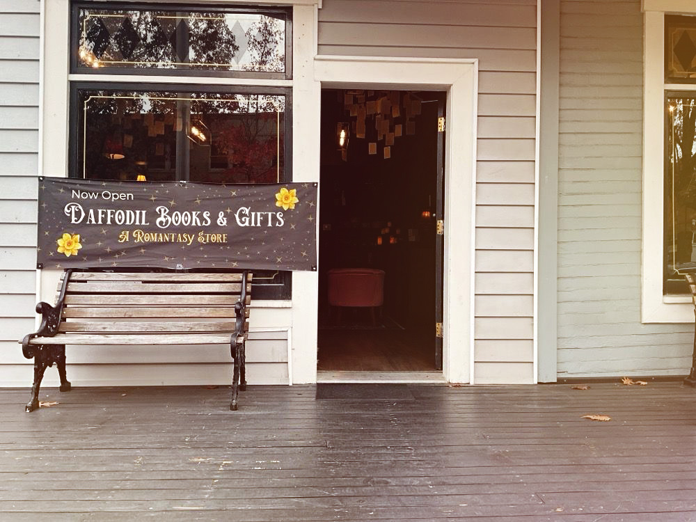 The outside of Daffodil Romantasy Books in Eugene, Oregon. It's a building with white siding. In this photo, there's a bench out front, and the door is open. You can see a chandelier with book pages hanging down from it inside.