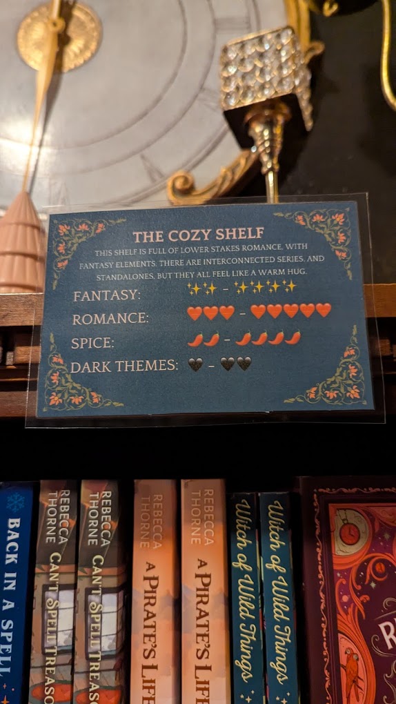 A sign on top of a bookshelf reads: The Cozy Shelf. It has a rating scale for the books on the shelf with symbols for fantasy, romance, spice level, and dark themes.