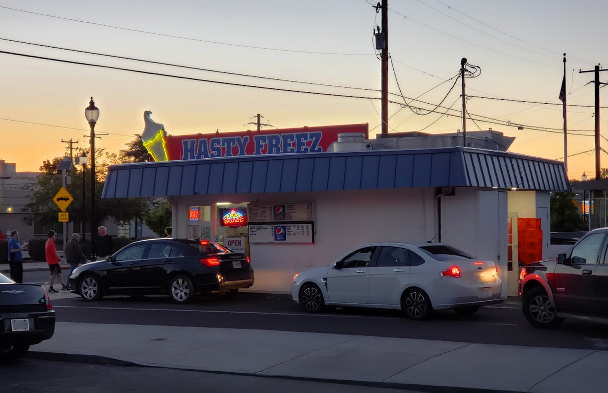 The outside of Hasty Freez at night time. There's a line of cars at the drive up window. It's a small white building with a blue roof, and a red sign with blue letters on top declaring 'Hasty Freez'. On the sign is a white and yellow neon ice cream cone.