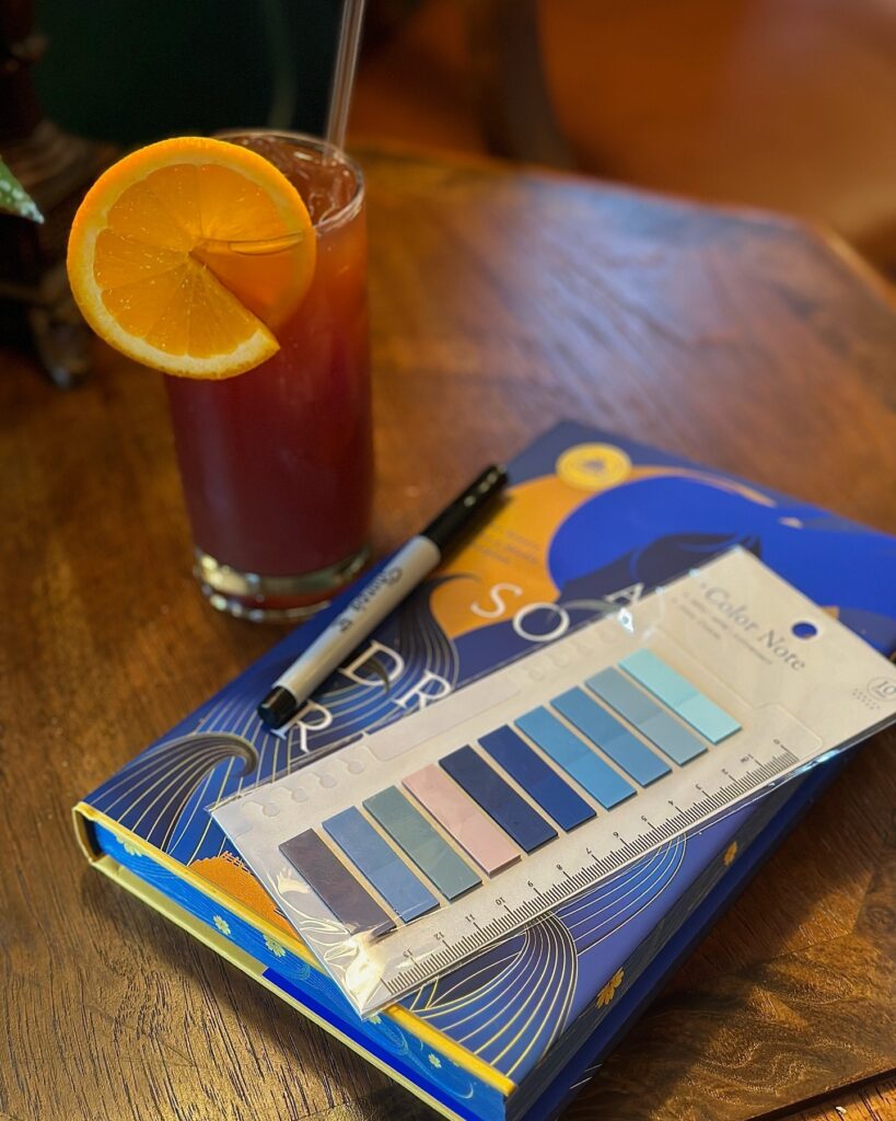 A blue book sits on a table next to a mocktail. There are also pretty blue color coded sticky notes and a black pen.