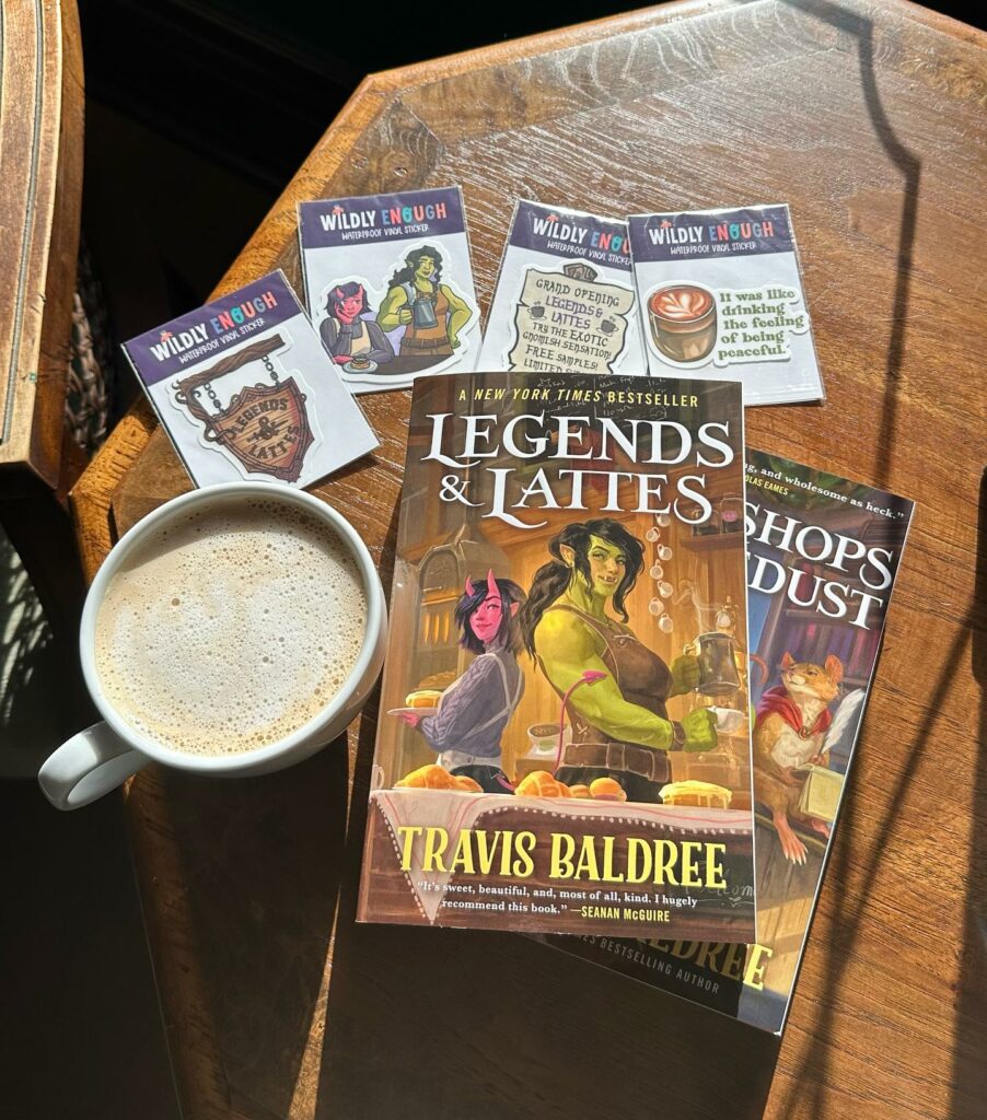 Two books, including Legends & Lattes, sit on a table next to a cup of coffee and stickers featuring things from the Legends & Lattes book inside of Daffodil Romantasy Books in Eugene, Oregon.