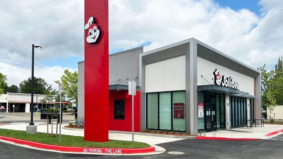 The exterior of a Jollibee restaurant. The building is boxy and modern looking, with gray paint, large windows, and splashes of bright red color.