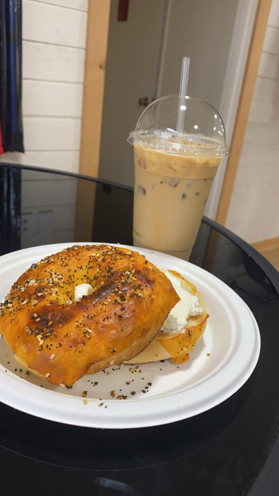 A bagel with cream cheese on a white plate, with a to-go cup of coffee behind it.