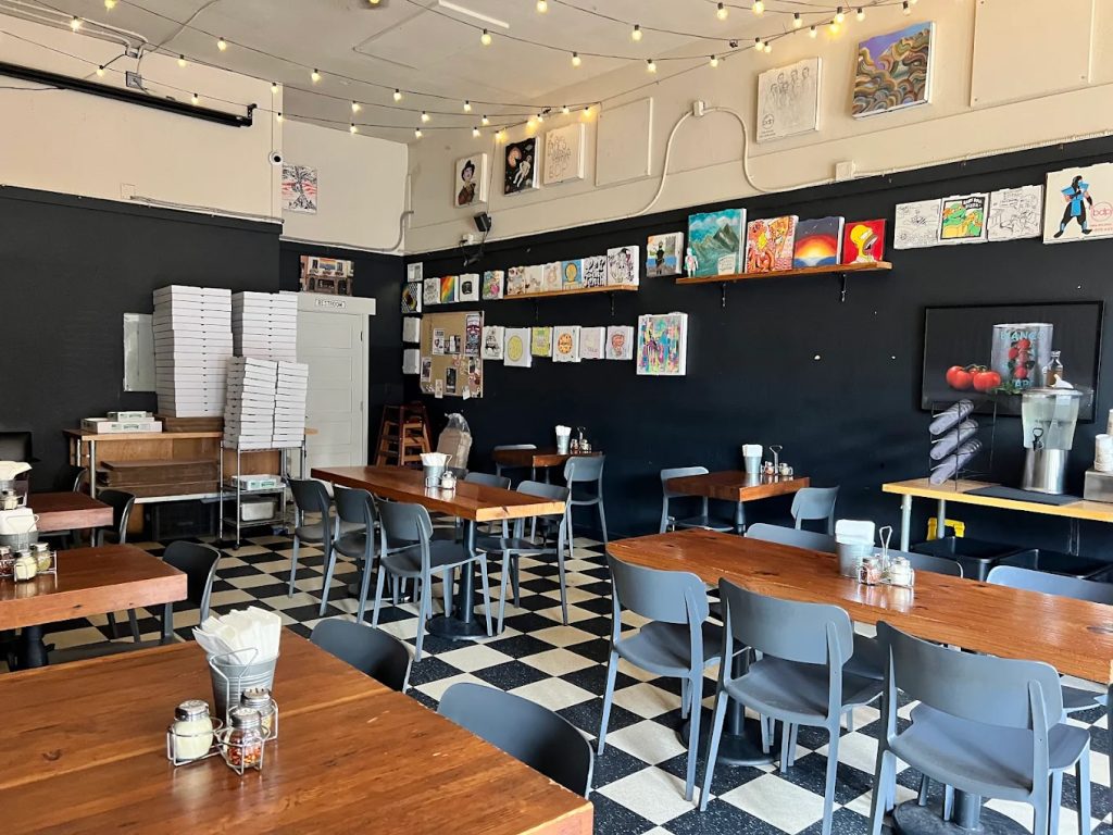The interior of Baby Doll Pizza. There are black and white checkered floors, black walls, lights strung across the ceiling, and eclectic art up on one wall. There are wooden tables and gray chairs.