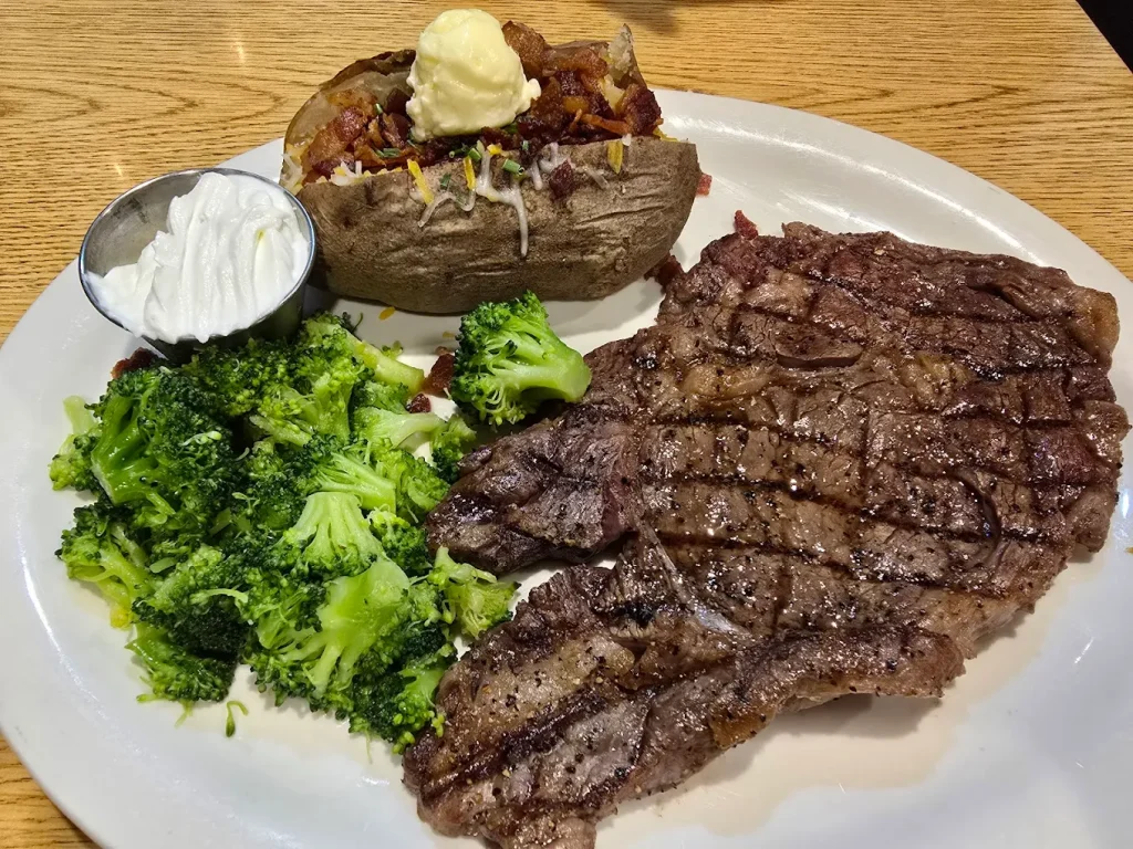 A huge steak with broccoli and a baked potato on a white plate.