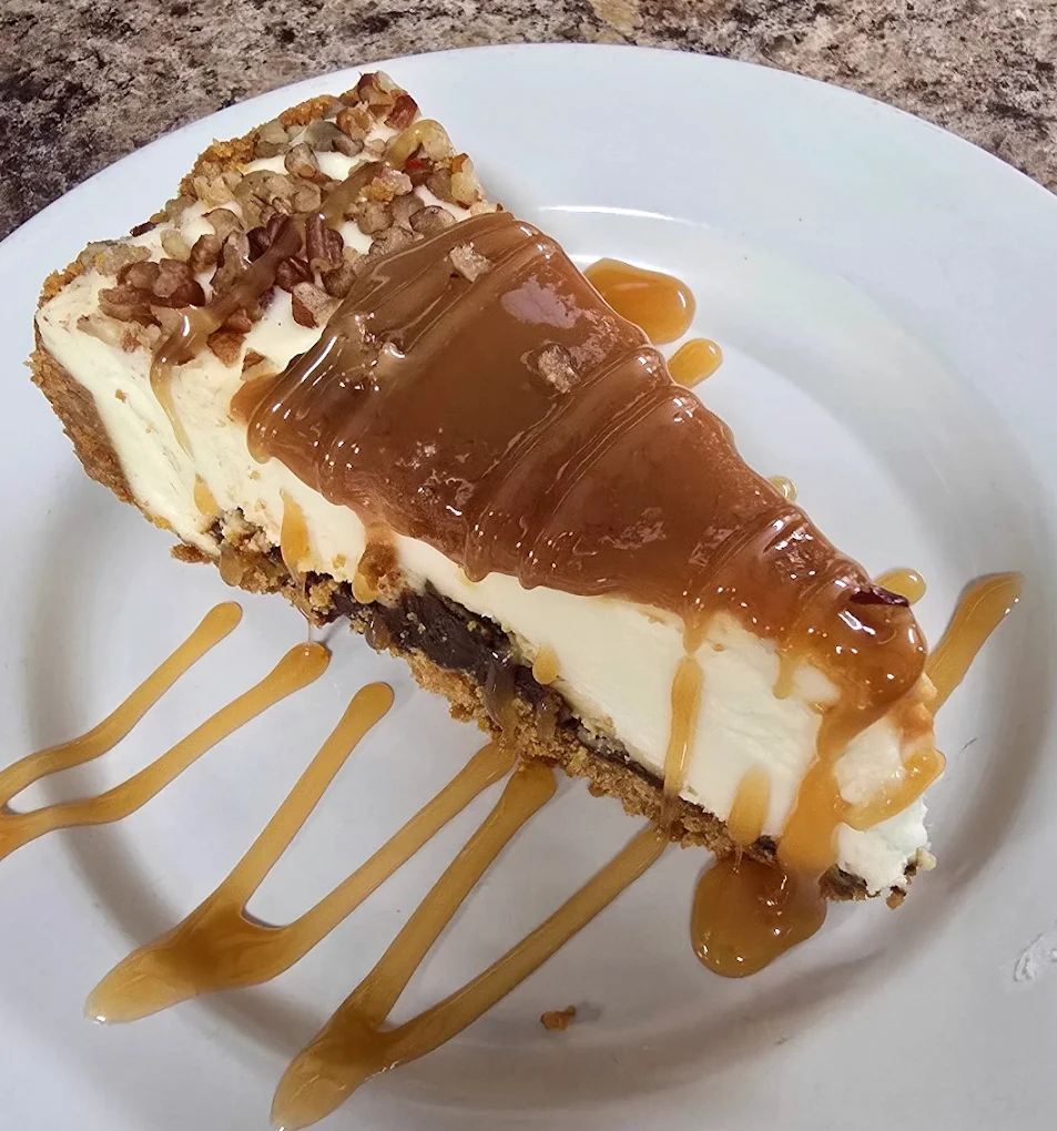 A piece of cheesecake covered in caramel and drizzled with even more caramel and topped with chopped nuts.