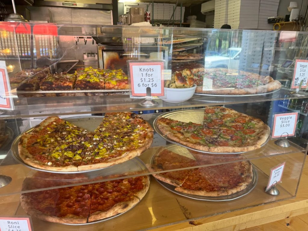 Pizzas sold by the slice sitting inside a glass display case at Baby Doll Pizza in Portland.