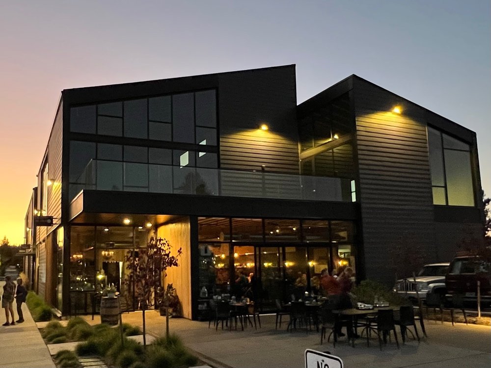 The outside of Rancher Butcher Chef in Bend, Oregon. It's a modern, sleek black building with a lot of windows and warm lighting outside. There are tables outside to eat at.
