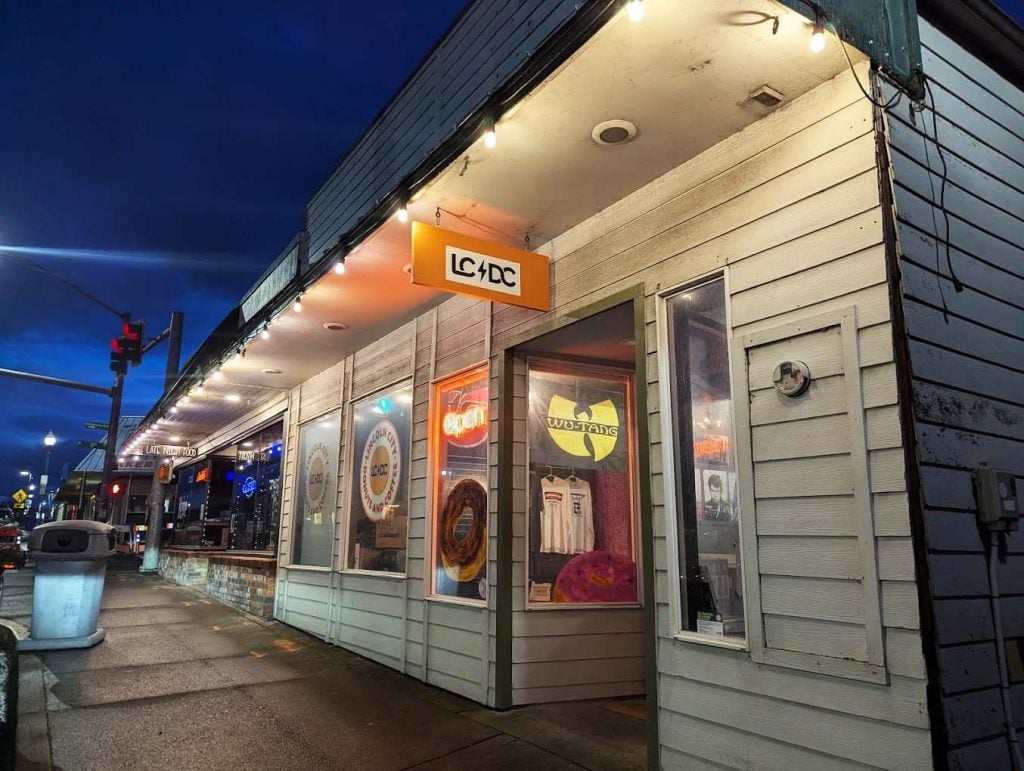 The outside of Lincoln City Donuts And Coffee. There are rock and other music band logos in the windows, and the sign for the donut shop says LC DC, a play on the band AC/DC.