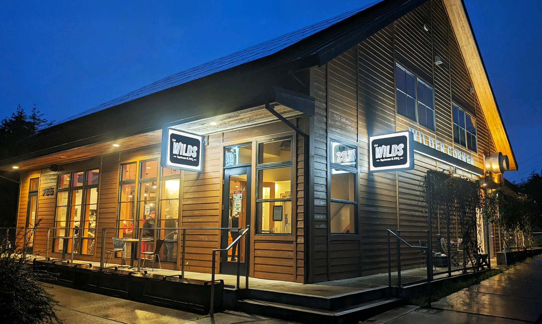 The outside of The Wild's Tap House & BBQ in Newport Oregon. It's a tall wood cabin style building, though modern. It had two black and white light up signs that say, 'The Wilds Taphouse & BBQ'. In the photo, it's night time, but a warm cozy glow emits from the windows and doors from inside the building.