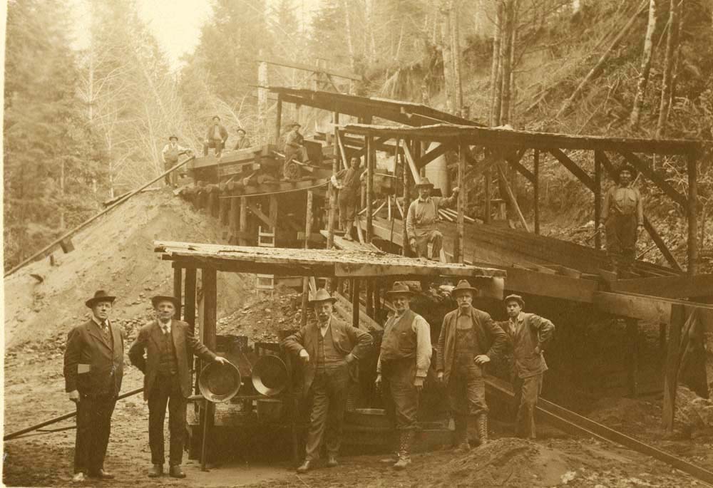 A historical photo of the Eagle Mine near Seven Devil's Road, around 1918. Men stand around a large piece of machinery. The photo is all in hues of brown.