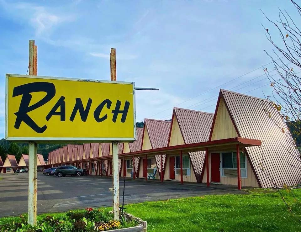 A bright yellow sign with black words declares 'RANCH'. The A-frame cabins stand in a row behind it. They are pale tan with a reddish, steeply sloped roof.