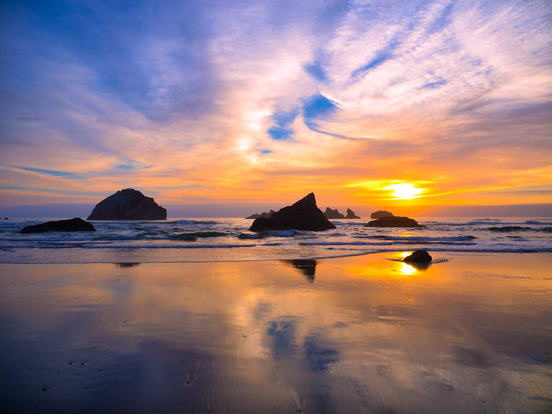 Rock formations rise up out of the surf near Bandon, Oregon at sunset. The sunset reflects off of the wet sand, and light airy clouds drift across the sky.
