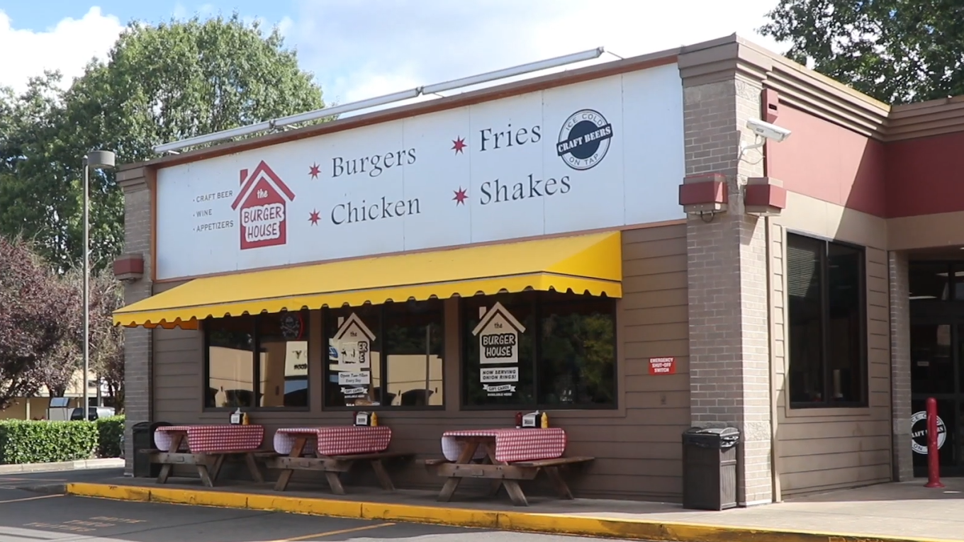 The outside of The Burger House. It's a brown brick building with a friendly yellow awning, and a large white sign with black words declaring: 'Burgers, Fries, Chicken, Shakes'.