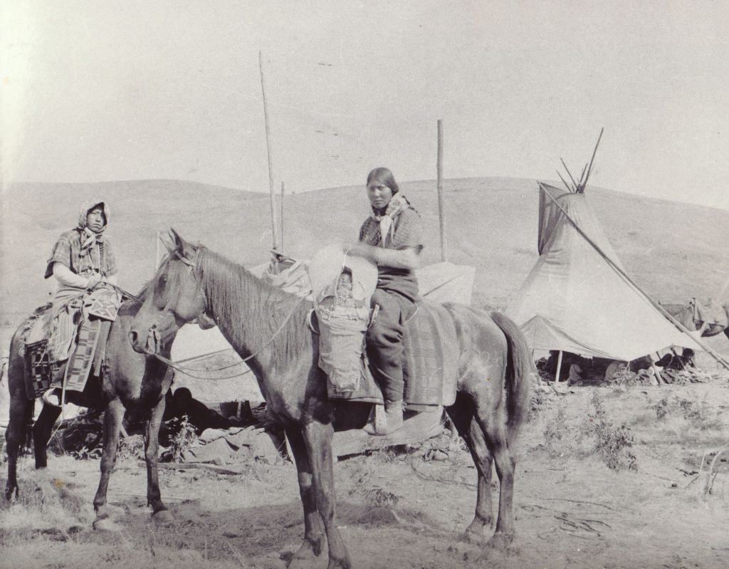 An old black and white historical photo of two Nez Perce sitting atop two horses.