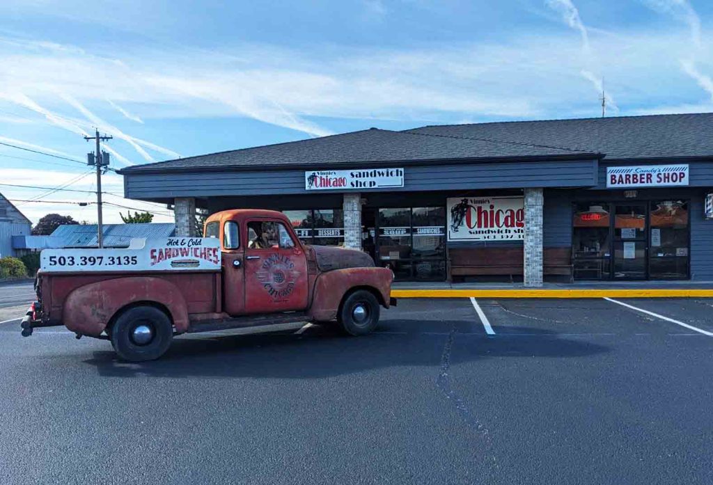 A vintage red truck sits in front of Vinnie's Chicago Sandwich Shop on a sunny day in St. Helen's Oregon.