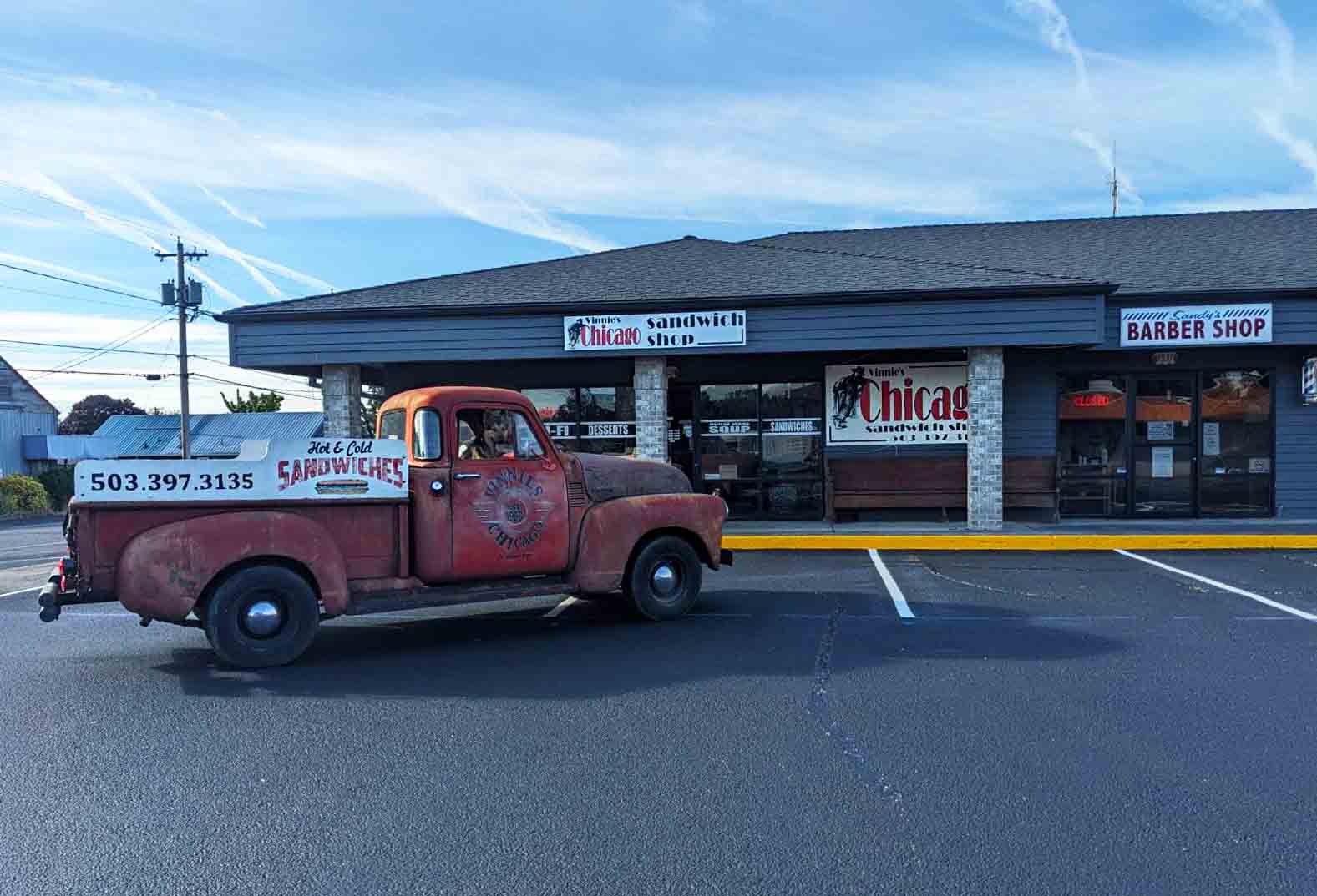 A vintage red truck sits in front of Vinnie's Chicago Sandwich Shop on a sunny day in St. Helen's Oregon.