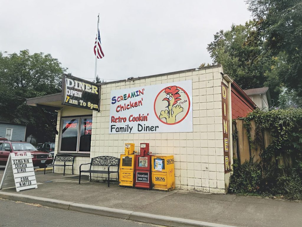 The outside of The Screamin' Chicken diner. It's a white building with a large sign of a screaming chicken. There are two black benches out in front on the sidewalk, and an American flag on a pole on the roof.