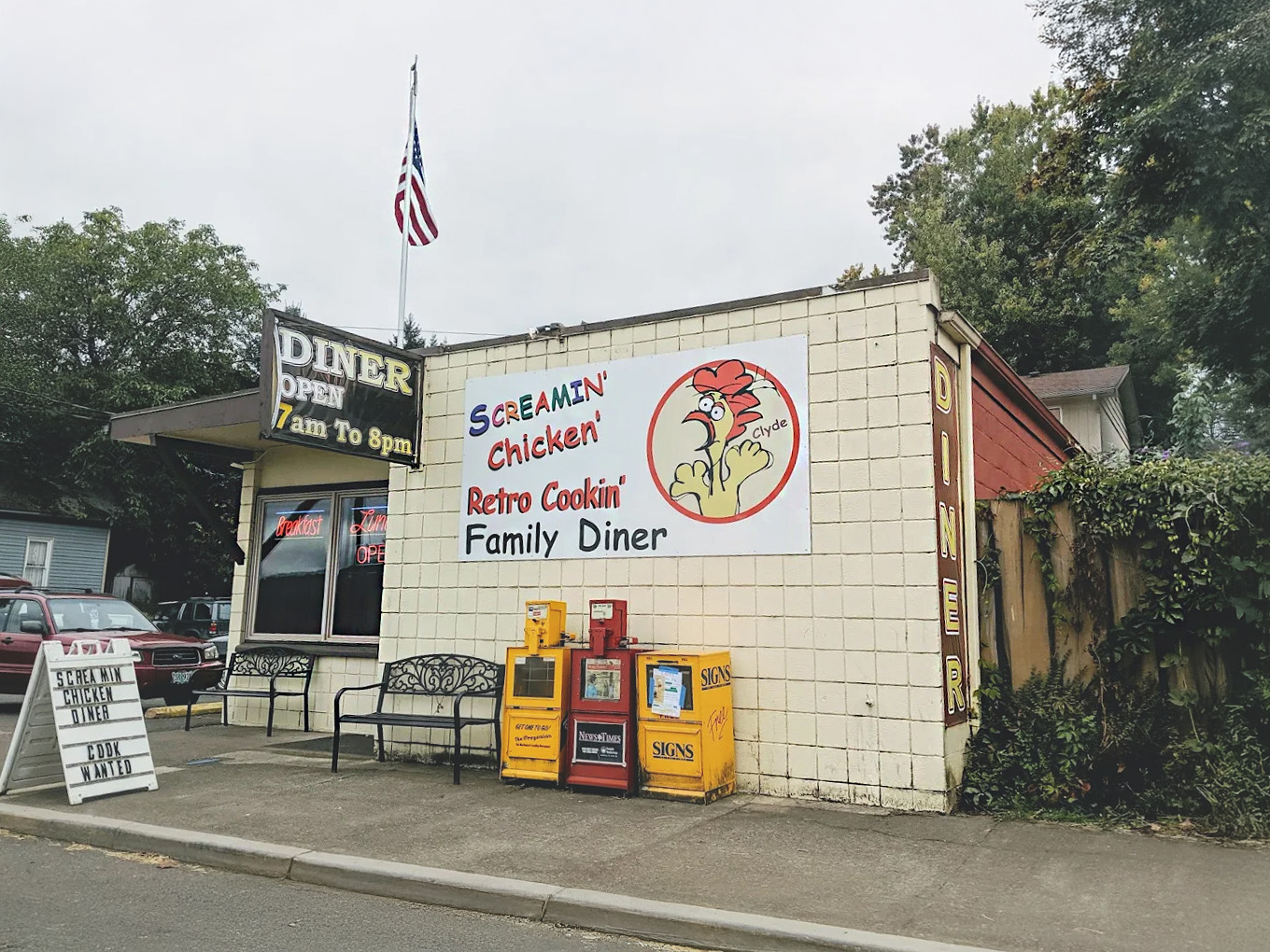 The outside of The Screamin' Chicken diner. It's a white building with a large sign of a screaming chicken. There are two black benches out in front on the sidewalk, and an American flag on a pole on the roof.