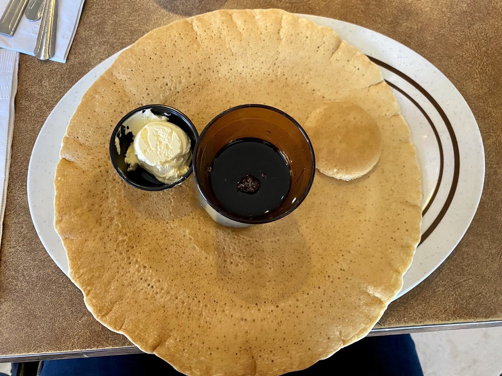 A massive pancake so big that it spills off the plate. It has a hole cut in the center of the pancake, and a cup of liquid sits inside the hole on the plate.