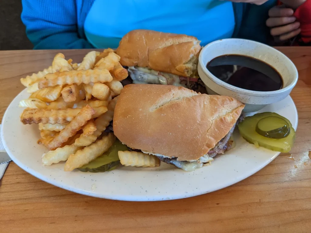 A large sandwich with dip, fries and pickles on a white plate.