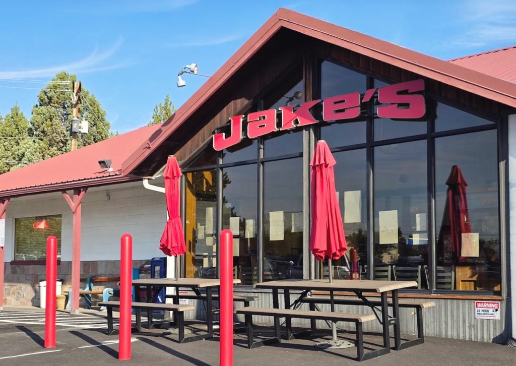The outside of Jake's Diner in Bend, Oregon. It's a white building with red words, a reddish roof, large front windows, and other red accents around the exterior.
