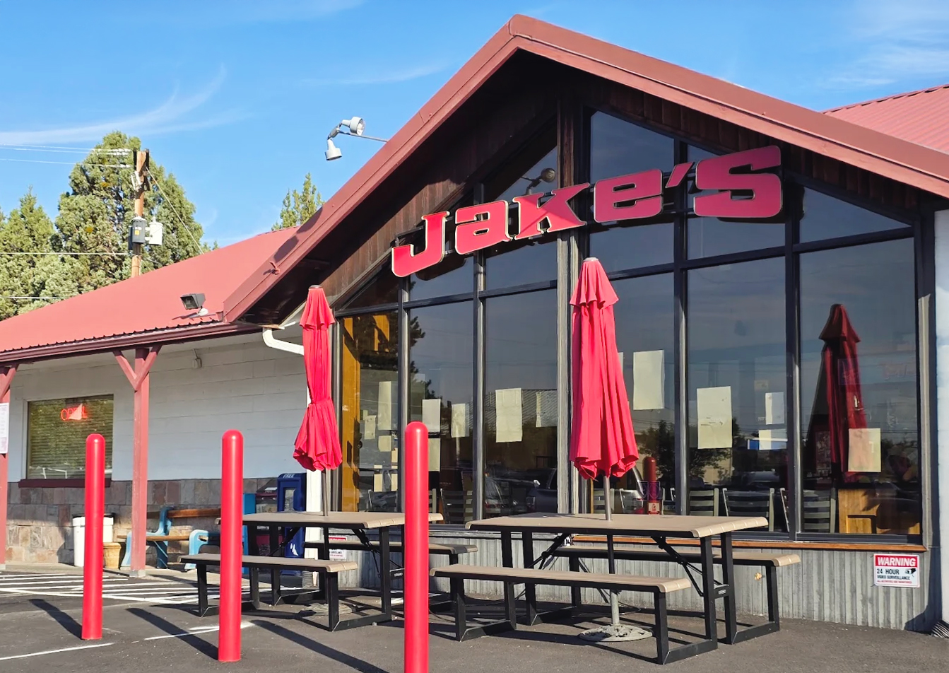 The outside of Jake's Diner in Bend, Oregon. It's a white building with red words, a reddish roof, large front windows, and other red accents around the exterior.