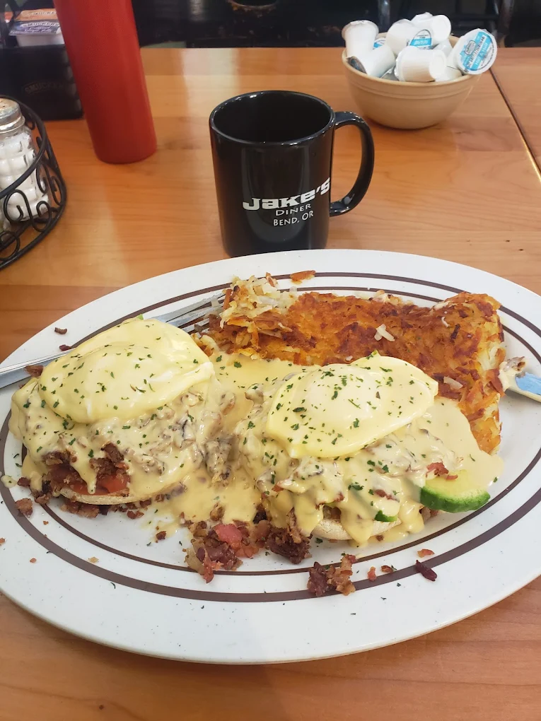 Eggs benedict with hollandaise sauce, avocado, bacon and hash browns. A black cup with the name 'Jake's' sits in the background on the table.