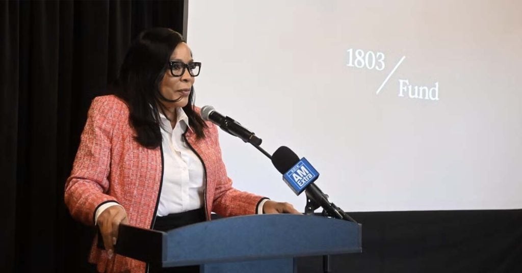 Portland Councilor Loretta Smith speaking at a podium with microphone during a public event, presentation slide visible behind her reading “1803 Fund”