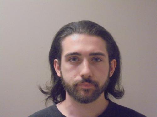 Booking photo of Anthony Trenkler, a white male with shoulder-length dark hair and facial hair, wearing a dark shirt, photographed against a plain background following his arrest.