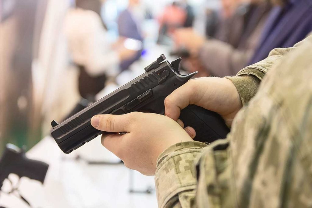 Close-up of a person holding and inspecting a semi-automatic handgun at what appears to be a gun store or firearms display, with other people blurred in the background.