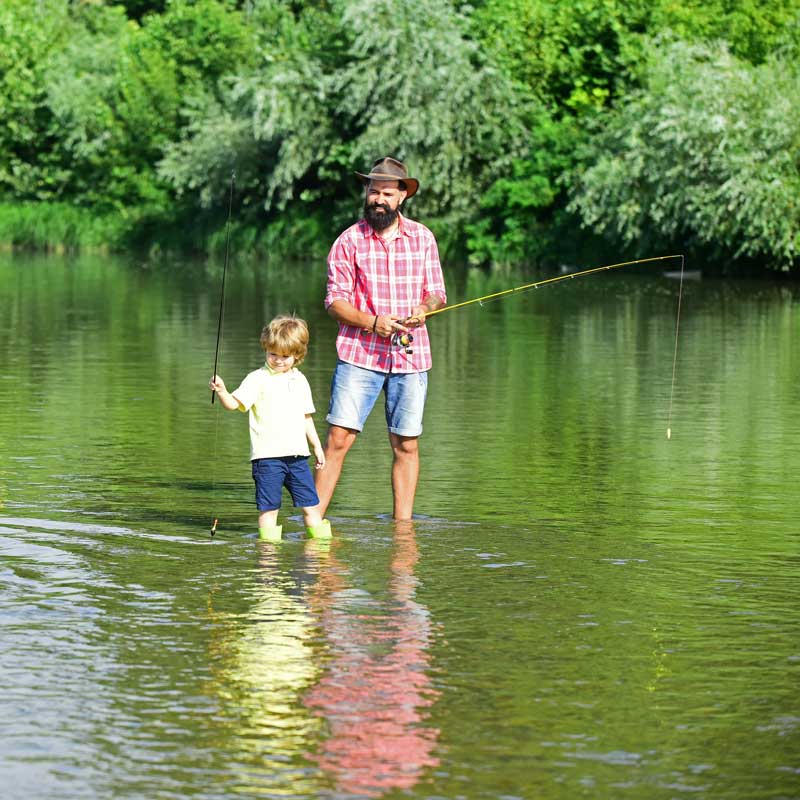Father and young child standing in a shallow river fishing together, surrounded by green trees and calm water in a natural outdoor setting