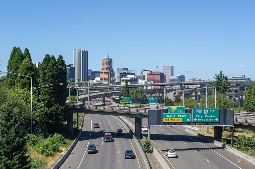 Downtown Portland skyline with Interstate 405 and Naito Parkway freeway signs in the foreground under a clear blue sky.