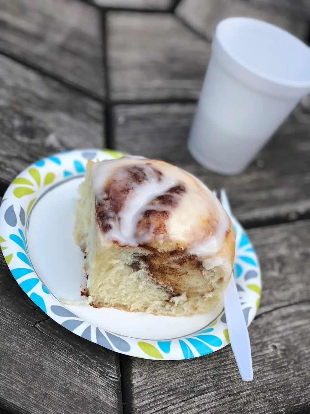 An iced cinnamon roll on a paper plate beside a cup on an outdoor wooden table at Country Bakery.
