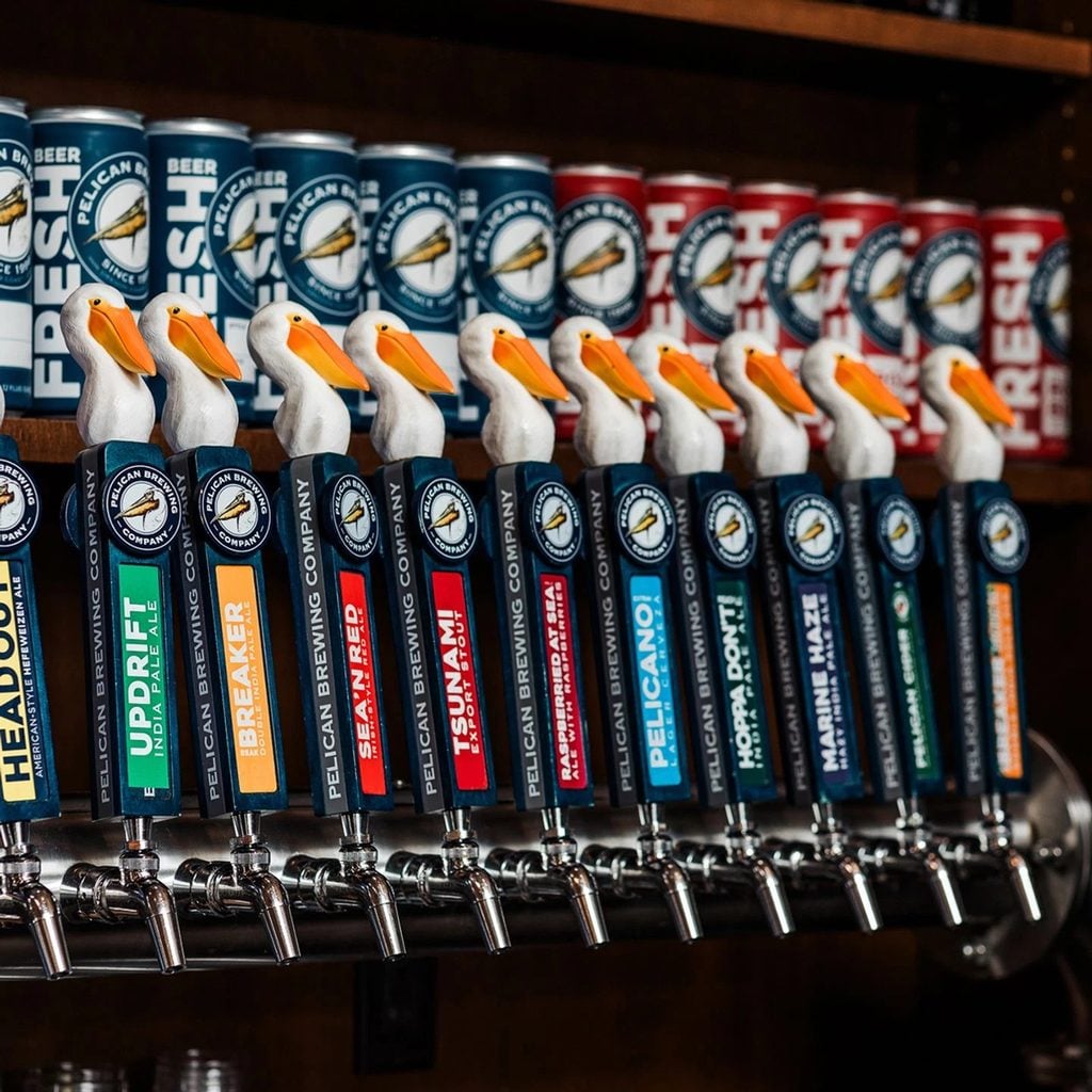 Row of Pelican Brewing beer taps topped with white pelicans with orange bills, with branded beer cans displayed behind them.