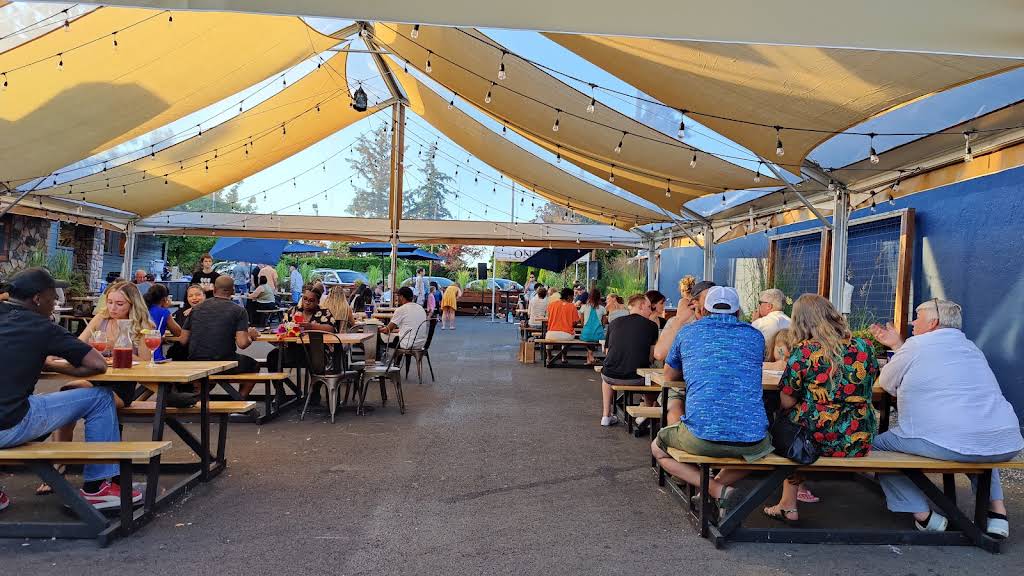 Diners at picnic tables beneath fabric shade sails and string lights on Amalfi’s Portland patio