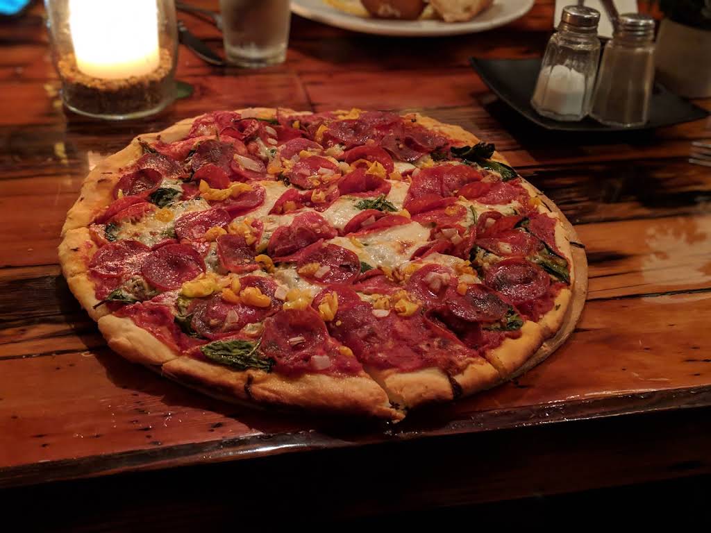 A whole pizza topped with sliced meat, peppers, and greens on a wooden table at Amalfi’s Italian Restaurant